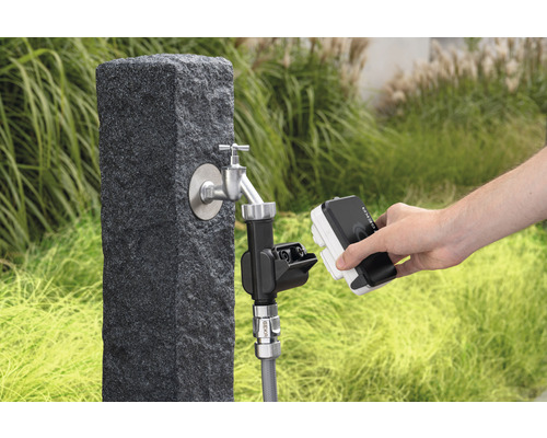 Gartenszene mit Wasserhahn an einem Steinpostament und einer Person, die ein Bewässerungsgerät anschließt