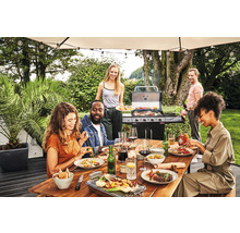 Gruppe von Freunden grillt und isst im Garten an einem Holztisch unter einem Sonnenschirm mit Grill