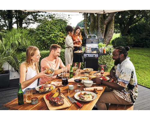 Eine Gruppe von Personen grillt und isst an einem Holztisch im Garten mit einem Gasgrill im Hintergrund.