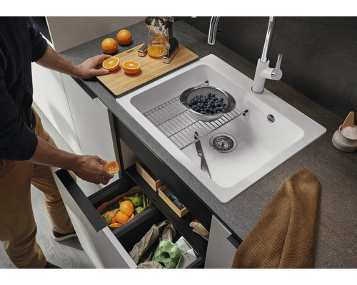 Küchenszene mit Spüle, Armatur, Schneidebrett, Orangen, Abfallbehälter und Person beim Entsorgen von Abfall.