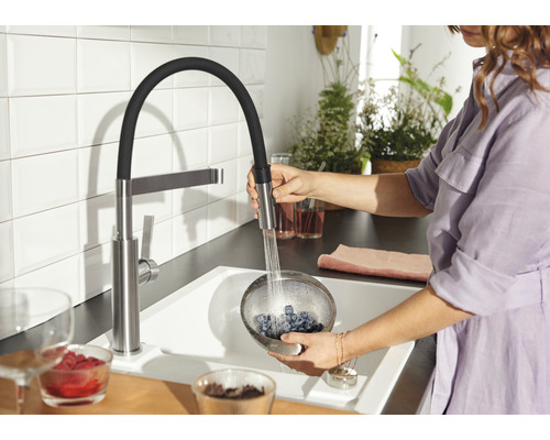 Frau wäscht Blaubeeren in einem Sieb unter einer Küchenarmatur mit ausziehbarer Brause in einer hellen Küche.