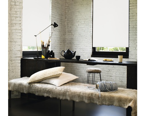 Heller Raum mit Rollos vor den Fenstern, einem langen Tisch als Arbeitsfläche und einer gemütlichen Sitzecke