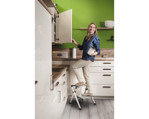 Frau auf einer Trittleiter in einer Küche mit weißen Schränken und Arbeitsplatten in Holzoptik