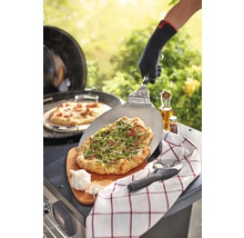 Eine Person bereitet im Freien Pizza auf einem Grill mit Pizzaschieber, Schneidebrett, Küchentuch und Knoblauch zu.