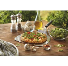 Szene mit Pizza auf Pizzaschieber, Knoblauch, Rucola, Öl, Gewürzen und Pizzaschneider auf einem Holztisch.