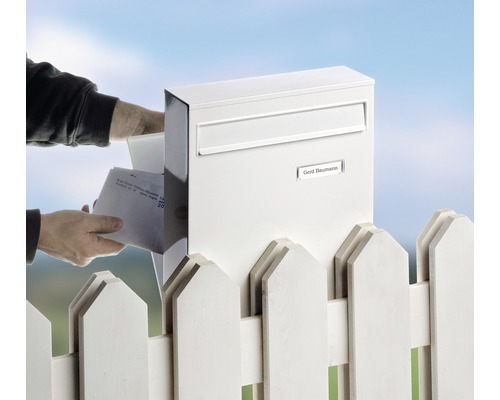 Person wirft Post in weißen Briefkasten neben weißem Zaun
