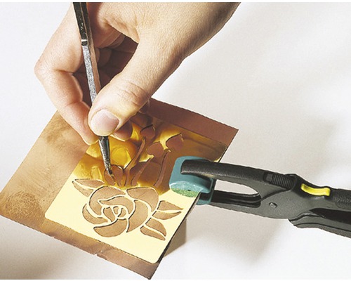 Eine Person graviert eine Rose in eine Metallplatte mit einem Gravierwerkzeug, die Platte wird von einer Klemme gehalten.