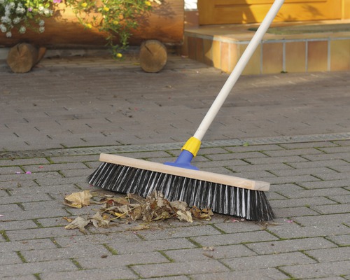 Kehrbesen mit Holzstiel und Borsten zum Reinigen von Laub auf einer gepflasterten Fläche.