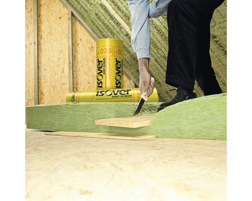 Ein Mann schneidet Dämmwolle von Isover in einem Dachboden zu. Dämmstoffrollen sind im Hintergrund zu sehen.