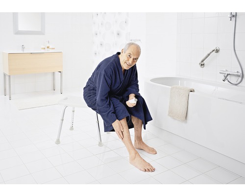 Mann sitzt auf einem Duschhocker in einem Badezimmer und cremt sich die Beine ein. Badewanne und Waschtisch sind im Hintergrund zu sehen.