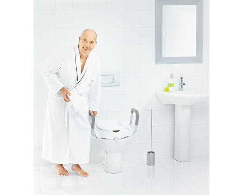 Ein Mann im Bademantel steht neben einer Toilette mit Toilettensitzerhöhung inklusive Haltegriffen.