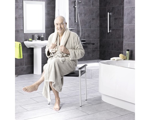 Ein Mann im Bademantel sitzt auf einem Duschstuhl in einem Badezimmer mit Dusche, Waschbecken und Badewanne.
