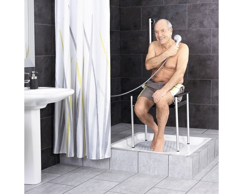 Badezimmer mit Duschvorhang, Waschbecken und Mann, der auf einem Duschstuhl in einer Dusche sitzt und eine Handbrause benutzt