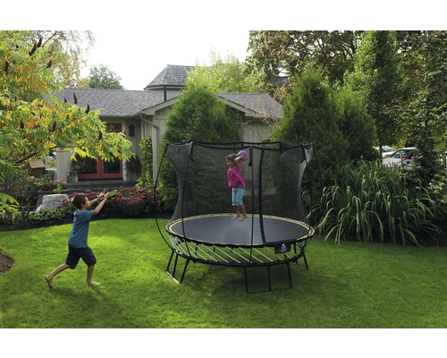 Kind springt auf einem Gartentrampolin mit Sicherheitsnetz, während ein anderes Kind den Ball fängt.