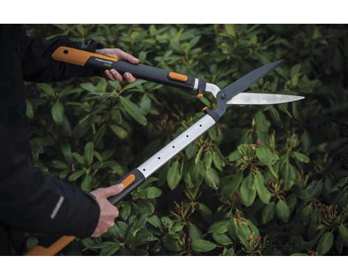 Eine Person benutzt eine Getriebe-Heckenschere vor einer grünen Hecke.