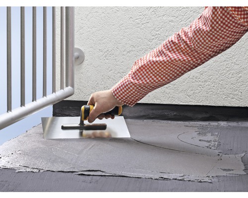 Eine Person glättet mit einer Glättkelle eine graue Beschichtung auf einem Balkonboden.