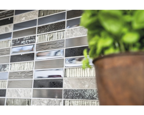 Detailaufnahme einer Wand mit Mosaikfliesen in verschiedenen Grautönen, neben einer Pflanze