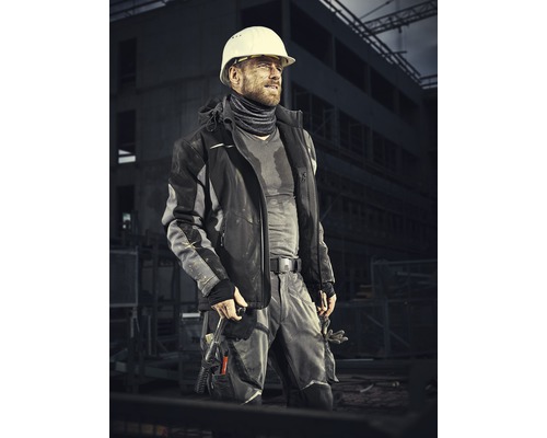Ein Bauarbeiter mit Schutzhelm trägt Arbeitskleidung vor einer Baustelle.