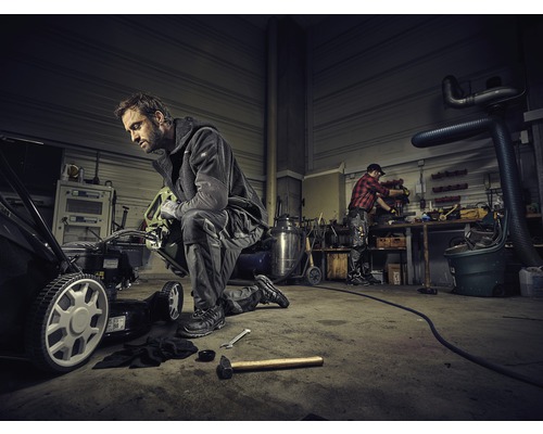 Werkstatt mit Mechaniker, der einen Rasenmäher repariert. Im Hintergrund arbeitet eine weitere Person an einer Werkbank.