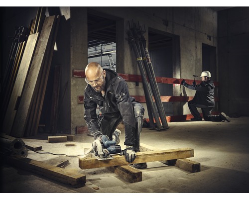 Ein Bauarbeiter sägt ein Holzbrett mit einer Kreissäge auf einer Baustelle. Im Hintergrund ist ein anderer Bauarbeiter zu sehen.