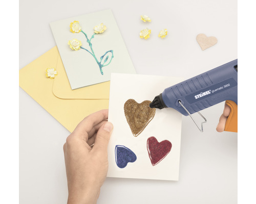 Hand, die mit einer Heißklebepistole Herzen auf eine Karte klebt. Bastelutensilien liegen daneben.