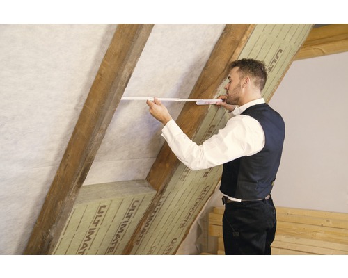 Ein Mann misst die Dachschräge mit einem Zollstock, wobei die Dachsparren mit Dämmmaterial verkleidet sind.