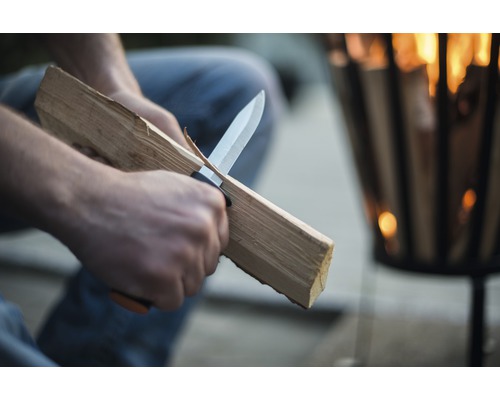 Eine Person schnitzt mit einem Messer Holz vor einer Feuerschale.