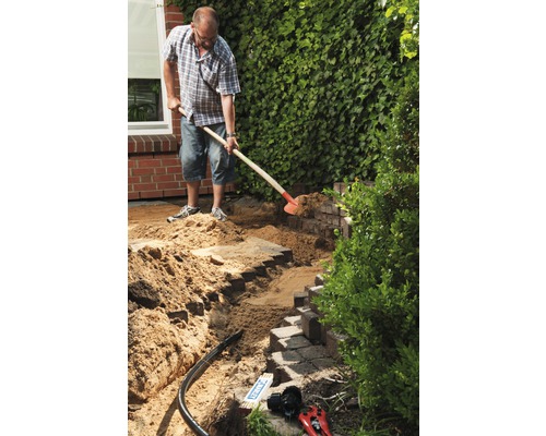 Mann arbeitet mit einer Schaufel im Garten an einer Mauer aus Pflastersteinen