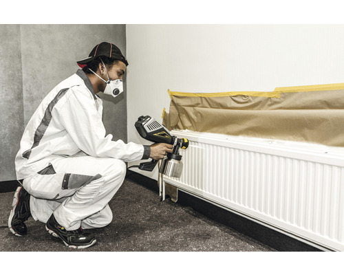 Eine Person lackiert mit einer Farbspritzpistole einen Heizkörper abgedeckt mit Malerkrepp und Papier.