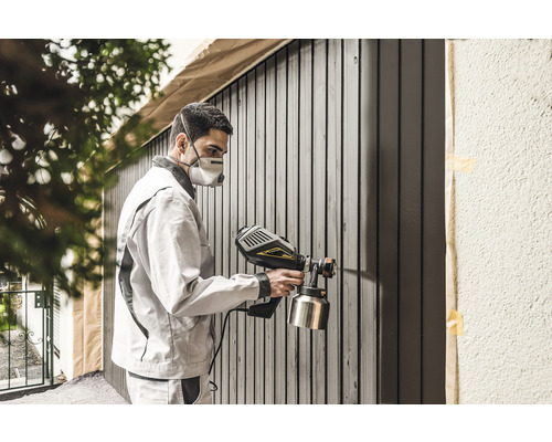 Mann mit Atemschutzmaske sprüht Farbe auf Holzzaun mit Farbsprühgerät