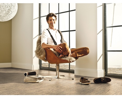 Ein Mann sitzt entspannt in einem Sessel in einem hellen Raum mit großem Fenster und Bodenbelag.