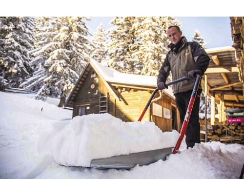 Mann schiebt Schnee mit einer Schneeschaufel vor einem Holzhaus.