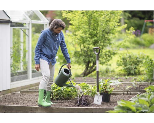 Frau gießt Pflanzen im Garten mit Gießkanne neben Spaten und Gewächshaus