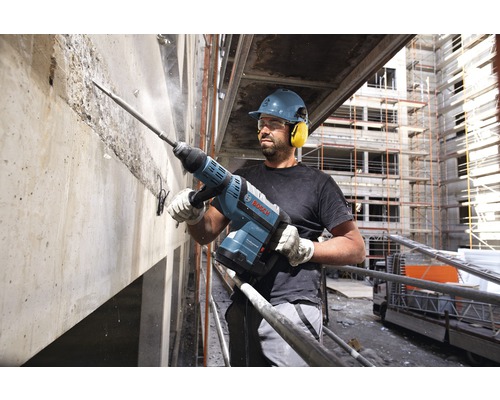 Ein Bauarbeiter mit Schutzhelm und Gehörschutz arbeitet mit einem Abbruchhammer an einer Betonwand. Bosch Logo.