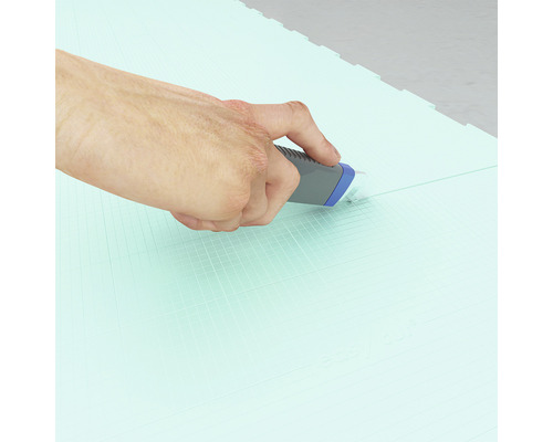 Eine Hand schneidet mit einem Universalmesser eine grüne Bodenmatte zu.