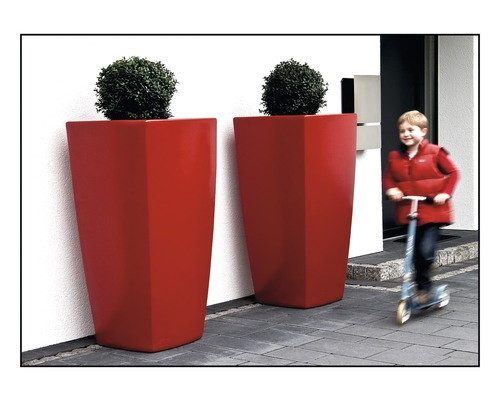 Zwei rote Pflanzkübel mit Pflanzen vor einem Haus, ein Kind fährt mit einem Roller vorbei.