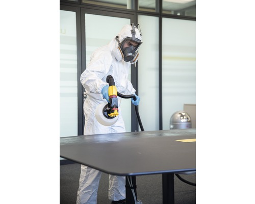 Eine Person in Schutzkleidung desinfiziert einen Tisch mit einem Desinfektionsmittel-Sprühgerät.