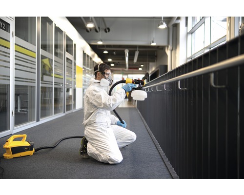 Mann im Schutzanzug desinfiziert mit einem Sprühgerät ein Geländer in einem Innenraum.