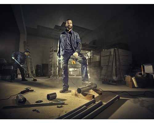 Handwerker in Arbeitskleidung mit Bohrmaschine in einem Lagerraum mit Rohren und Werkzeugen.