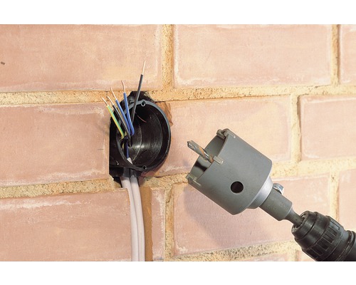 Bohrmaschine mit Bohrkrone neben einer Wandsteckdose in einer Backsteinmauer