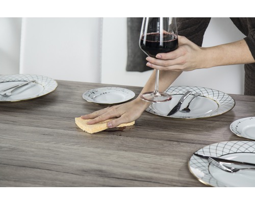 Szene mit Tisch, Gedeck und einer Person, die einen Fleck mit einem Schwamm aufwischt, während sie ein Weinglas hält.