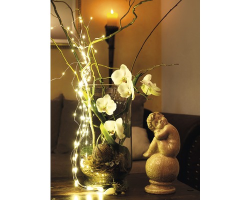 Dekorative Glasvase mit Orchideenblüten, Zweigen und Lichterkette neben einer Engelstatue