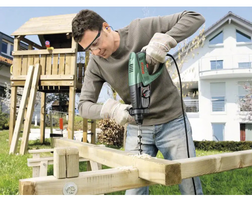 Ein Mann bohrt mit einer Schlagbohrmaschine Löcher in Holzbalken im Garten, im Hintergrund ein Spielturm.