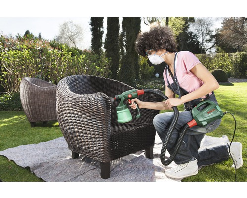 Frau sprüht mit einer Farbspritzpistole einen Korbstuhl im Garten
