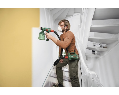 Mann streicht Wand mit Farbsprühsystem in Treppenhaus, Schutzfolie
