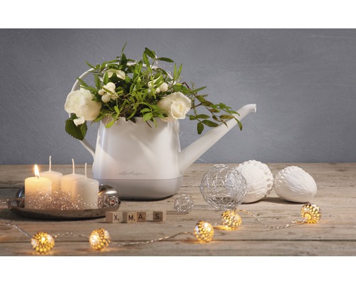 Dekorative Gießkanne mit Blumenschmuck, Kerzen, Kugeln und Lichterkette auf einem Holztisch