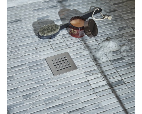 Detailaufnahme einer Dusche mit Bodenfliesen, Duschabfluss, Bürste, Badezusatz und Seifenschaum