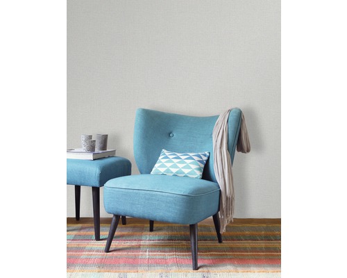 Innenraumszene mit hellgrauer Tapete, türkisfarbenem Sessel, Hocker und buntem Teppich