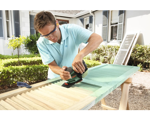 Ein Mann verwendet einen Schleifer, um eine Holzjalousie zu bearbeiten. Die Jalousie liegt auf einem Holzbock.