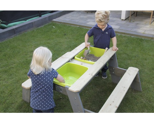 Zwei Kinder spielen an einem grauen Sand- und Wassertisch mit zwei grünen Wannen im Garten.